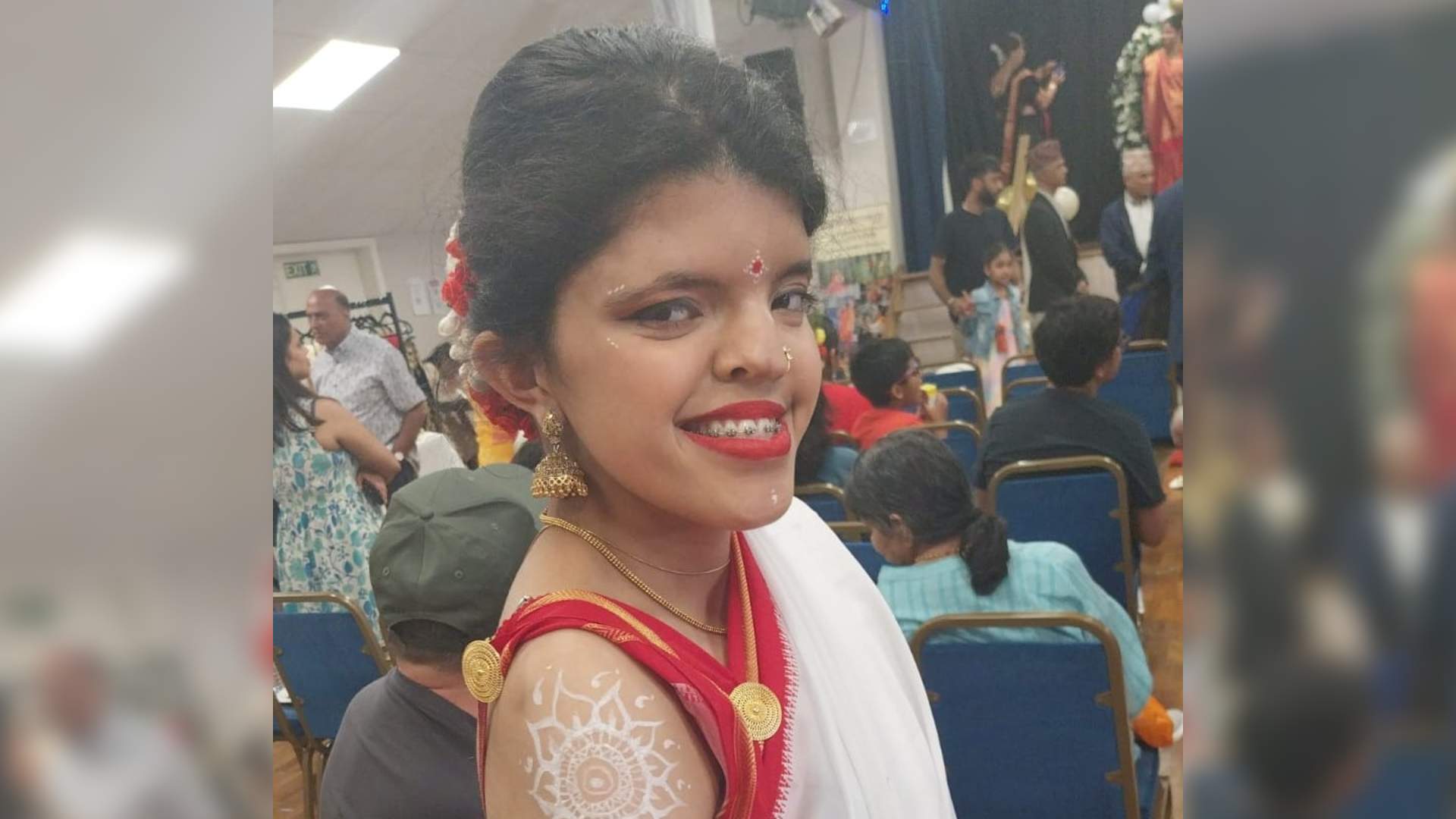 Sneha is a south Asian woman. She has craniofacial dysplasia. Sneha wears a red sari and red lipstick.