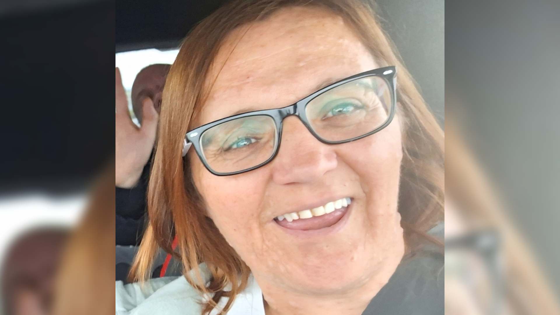 Hayley has neurofibromatosis. She takes a selfie inside a car. Hayley wears rectangular black glasses and smiles widely at the camera.