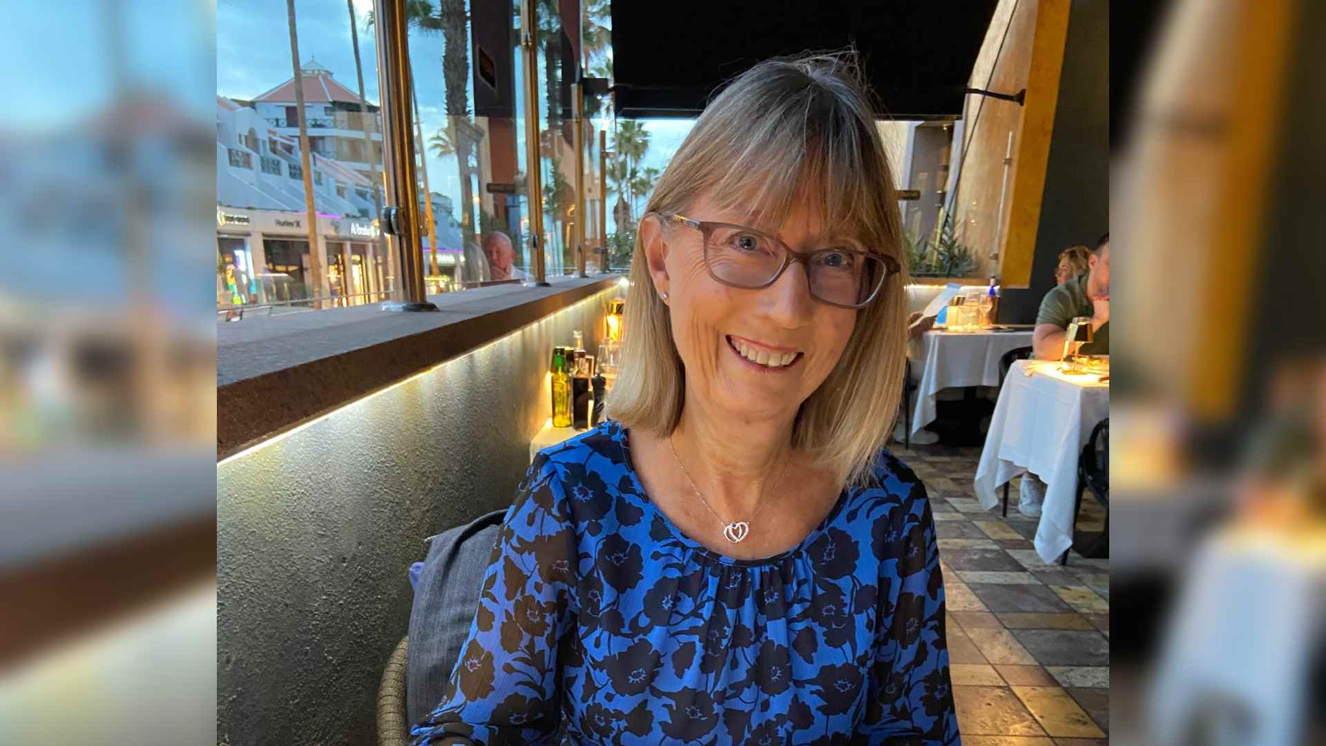 Janelle has Juvenile Dermatomyositis causing a telangiectasia rash on her face. She sits in a restaurant wearing a blue patterned top. She smiles at the camera.