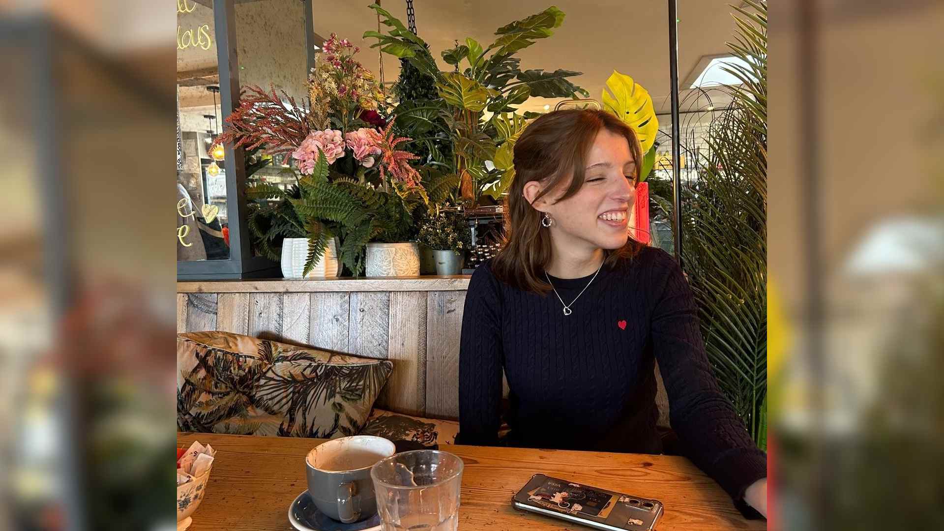 Rose sits in a cafe with a variety of plants behind her. She looks off to the side smiling. Rose has naevus anaemicus and scarring following the removal of haemangioma birthmarks.