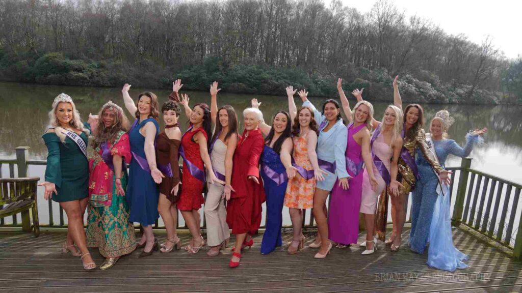 A photo of a group of women posing outside by a lake. They are all wearing pageant outfits in various bright colours.