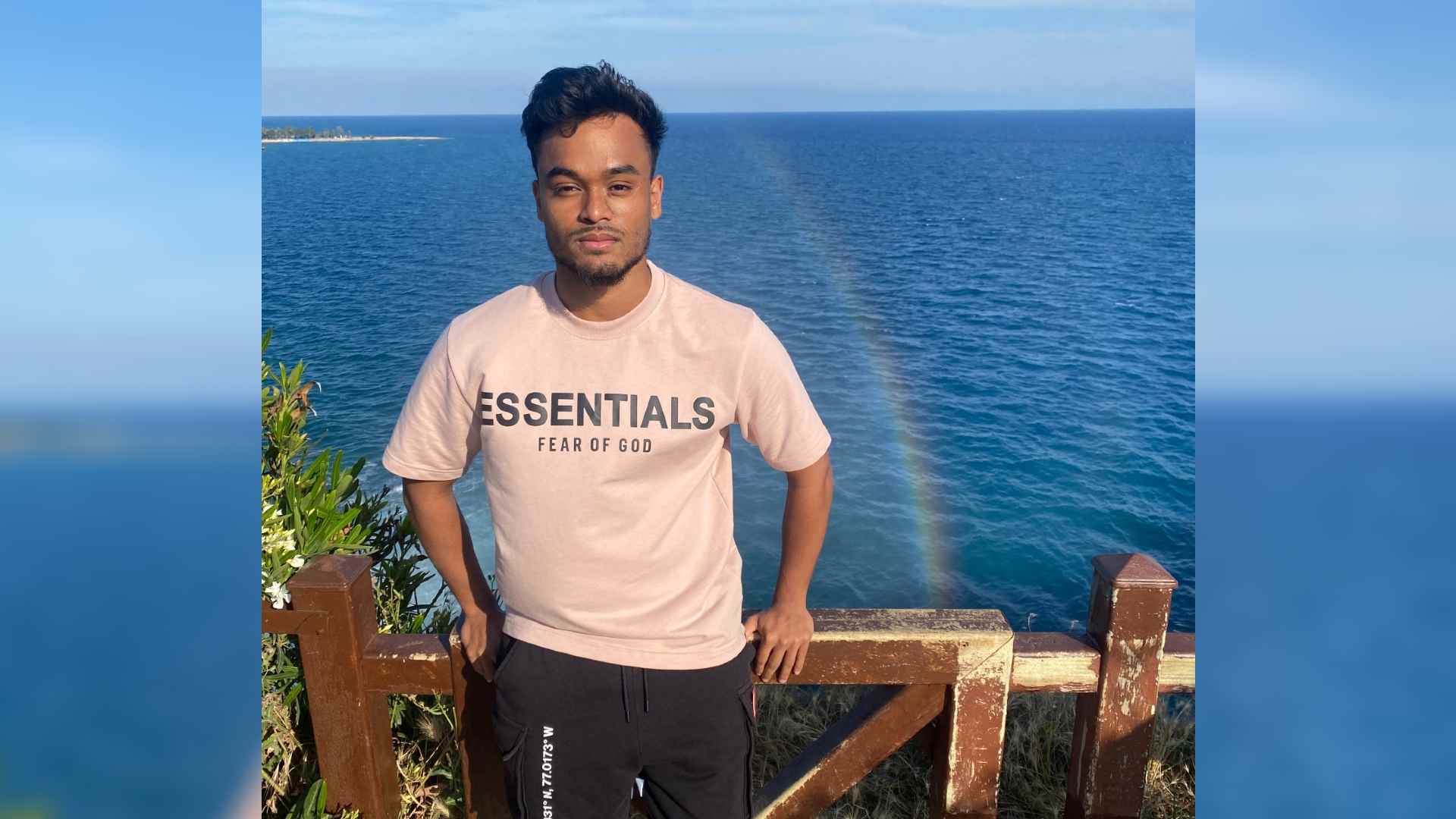 Yunus has facial scarring following the removal of a birthmark. He stands leaning against a fence with the sea behind him.