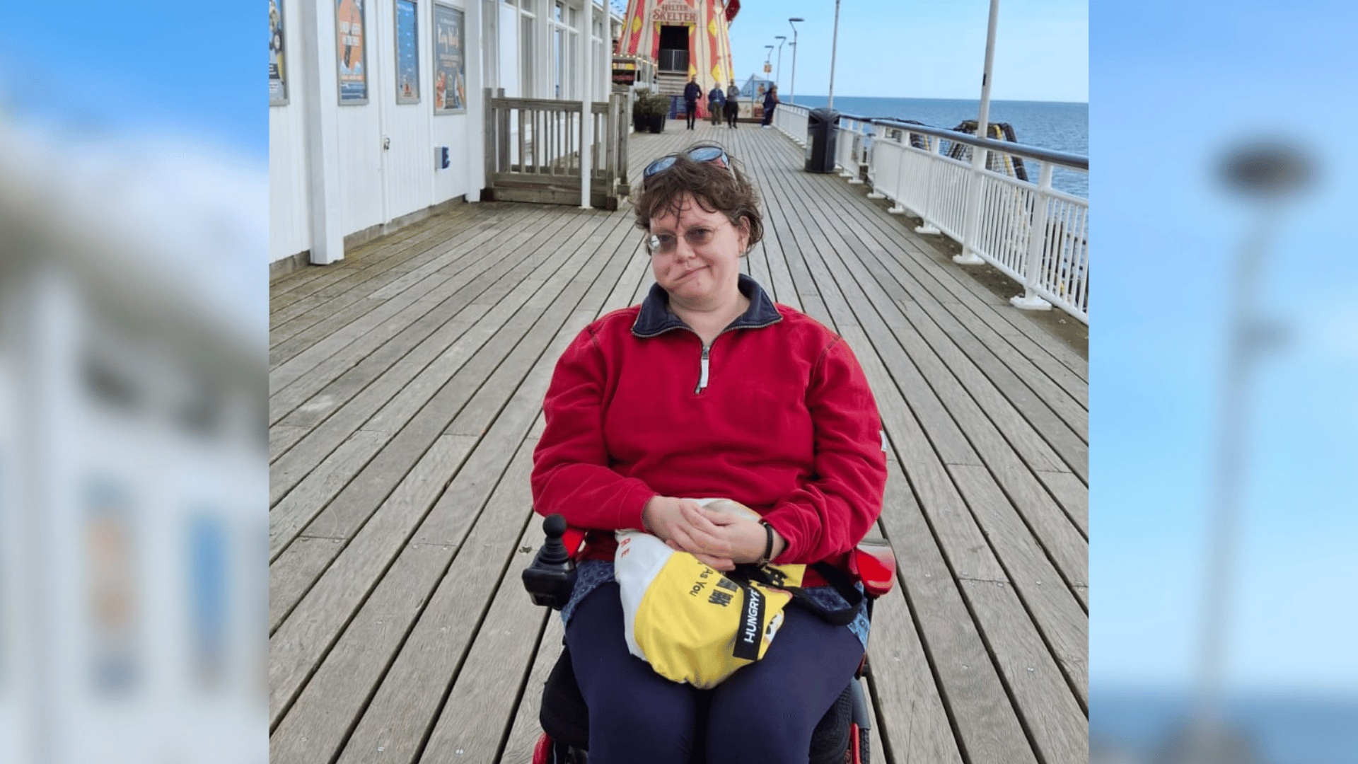 Angela sits in a wheelchair on a pier. A helterskelter can be seen in the background. Angela has facial palsy.